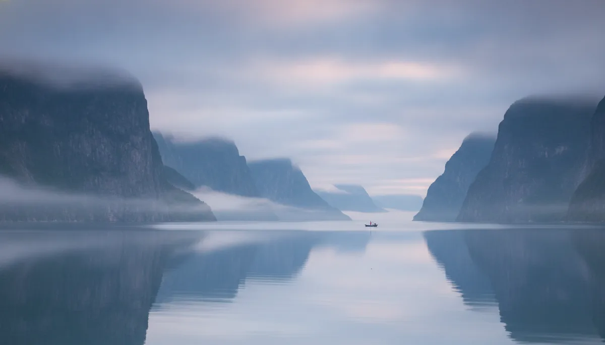 Misty fjord at dawn, low clouds between steep cliffs, calm water, muted blues and greys, wide cinematic shot